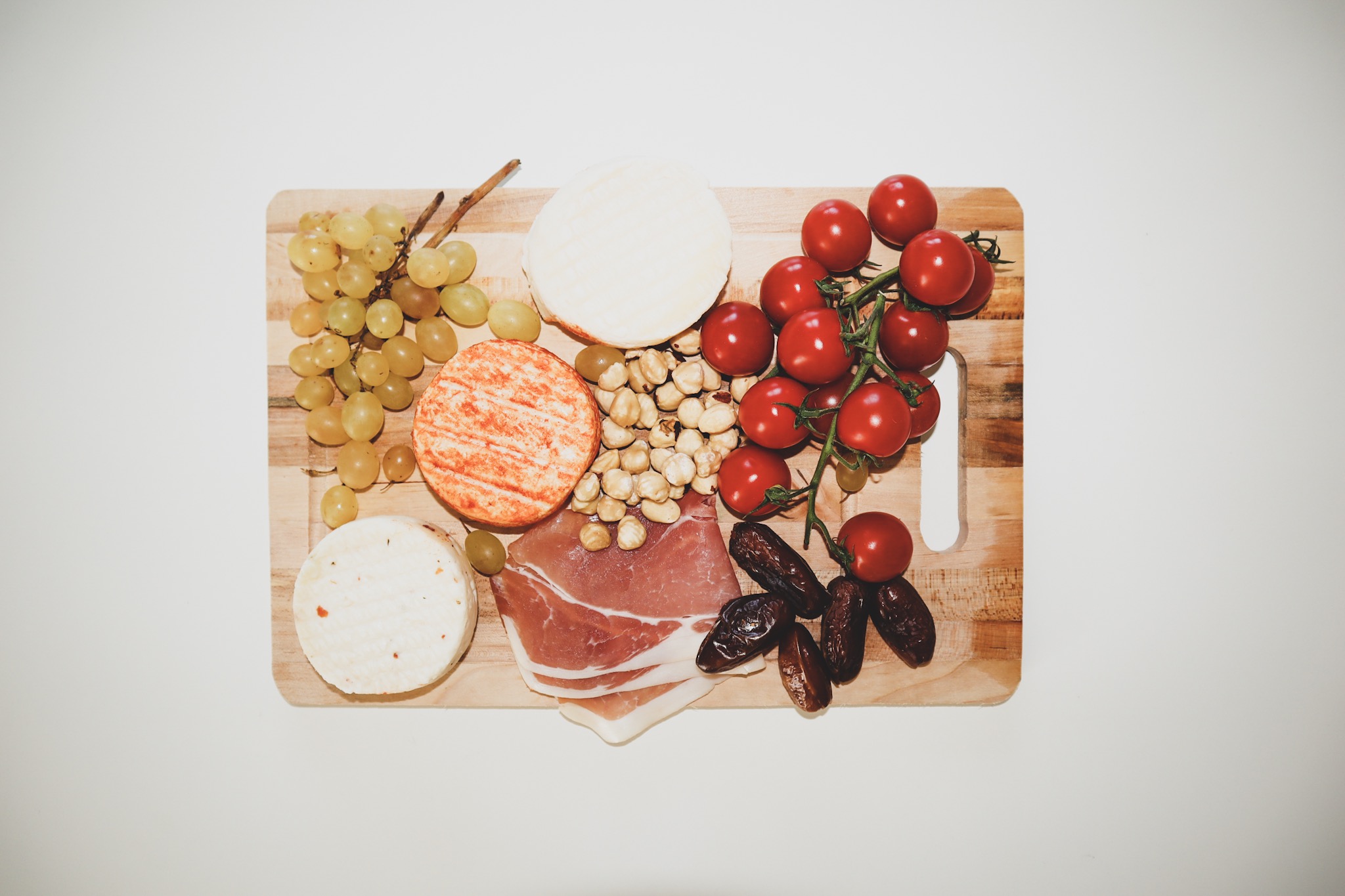 a wooden tray filled with grapes, prosciutto, cheeses, figs, and cherry tomatoes against a white backdrop