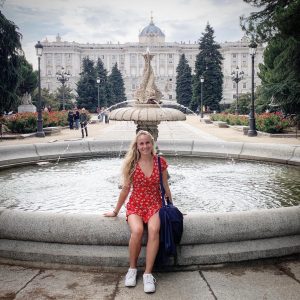 A girl in a bright red dress sits on the edge of a fountain in front of a large European palace.