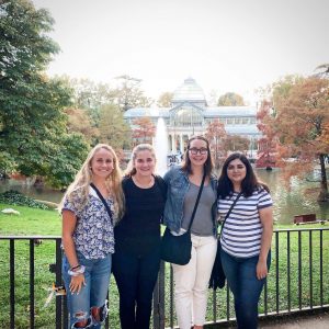 Four women stand in front of a grassy field on a cloudy day. There is a European palace and fountain in the background.