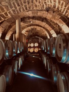 A wine cellar with stone/brick ceilings and arches, with many barrels of wine lining both edges of the walkway