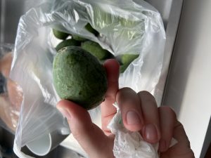 A small green oblong shaped fruit being held by a hand over a bag of more fruit