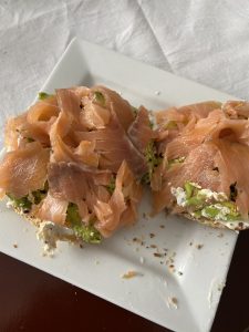 A bagel with avocado, cream cheese, and smoked salmon on a white plate on a white tablecloth