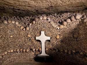 A cross tombstone surrounded by bits of human skulls and bones in arch shapes in the cave of the catacombs.
