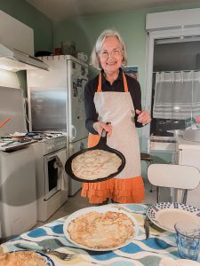 73 year old woman wearing a white and orange apron holding out a skillet with a freshly made crêpe.