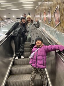 4 people facing the camera riding down an escalator, a dad, and two young women and then a daughter at the bottom.