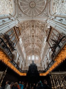 Gold accented carved white stone ceiling of a cathedral with two organ pianos on either side.
