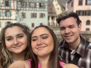 2 girls and 1 guy from left to right taking a selfie smiling with mouths closed and blurred out colorful buildings behind them.