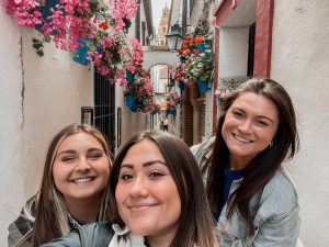 3 girls posed in a tiny alley of Spain with white walls and pink, and red flowers in blue vases hanging