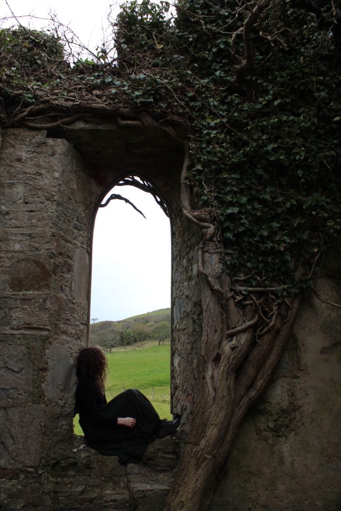 girl perched on a castle window with a tree branching around it looking out on green fields 