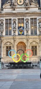 The olympic rings statue stand strong and illuminated in front of the large palace like structure that is town hall. 