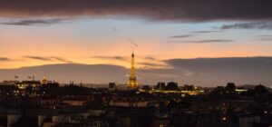 A sunset skyscape of blues, pinks, and greys with the Eiffel tower lit up at its center and the roofs of buildings spanning the bottom third of the photo