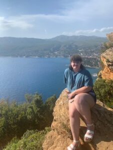 A photo of myself with the Mediterranean Sea and La Ciotat in the background. I am sitting on a cliff with trees and brown rocks below me 