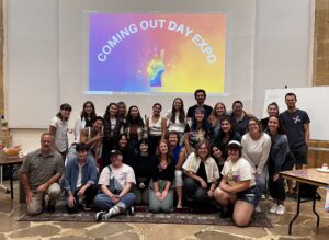 All attendees of the Coming Out Day Expo, allies and community members alike. Everyone stands/crouches in front of the backwall where the Coming Out Day Expo poster is projected in all of it's rainbow glory.