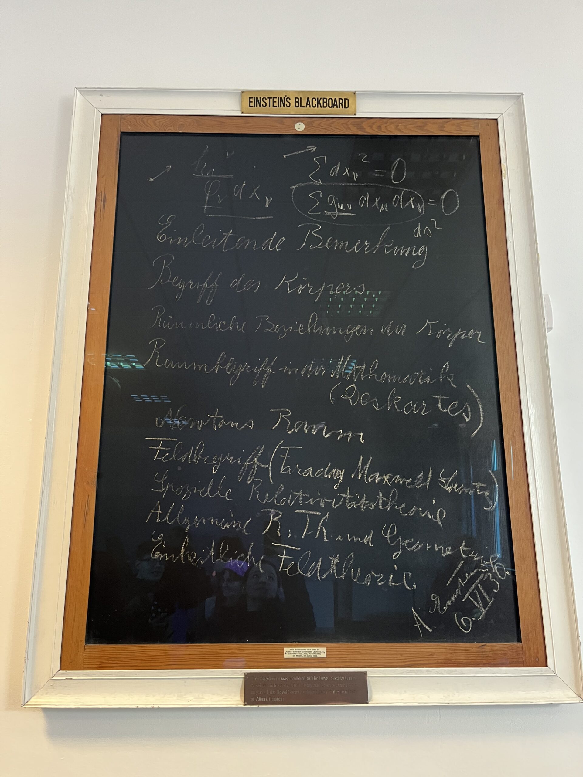 A cut out and framed piece of blackboard that Einstein wrote on. The frame is white and says “Einstein’s Blackboard” in black on a gold little plaque on the top. The blackboard is written on with white chalk. There is some equations written on the top of the Blackboard and the rest of the blackboard is words written in German. 