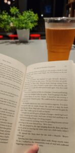 A book fills the bottom two thirds of the photo, with a grey table taking up the top third. On top are a plastic cup of cider and a fake potted plant.