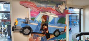 A large lego mural within the Lego store of Harry and Ron within the flying blue car from the second movie. Hedwig the white snow owl flies above them among the bright pink clouds