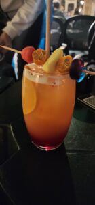 A cocktail sits in the center of the frame, its coloration fading from light orange to dark red. An orange is submerged within it and a wooden skewer of candies and small kumquats lays across the top. A straw sticks straight up behind it.