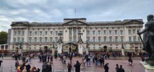 The beige-grey stone Buckingham palace stands tall above the metal and stone gates encompassing it. Tourists and locals alike stand in awe at the structure from the brick plaza in front of it all. Grey clouds float overhead.