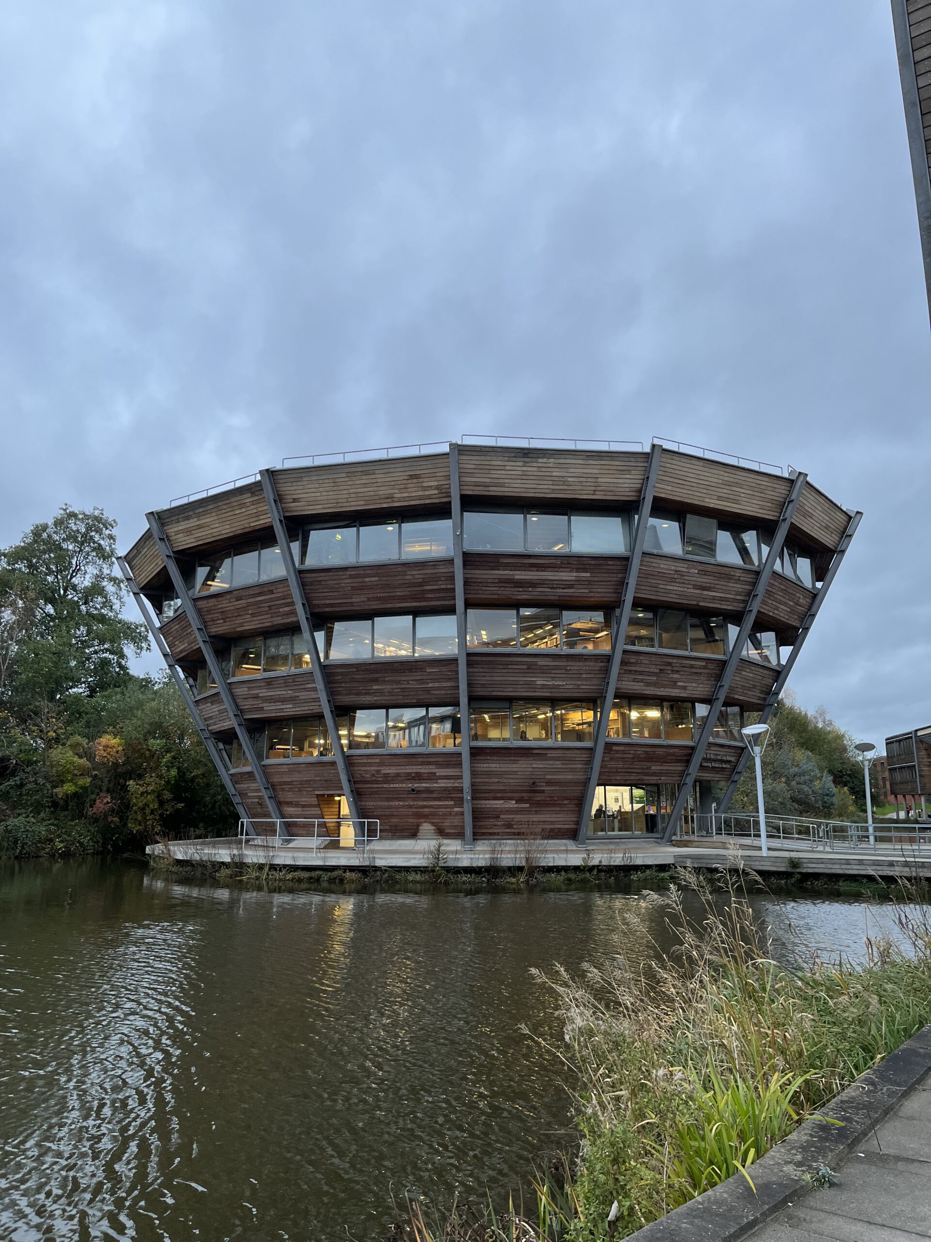 Displayed is a brown wood and lots of glass windowed bowl shape building with thin, steel, vertical beams. The building is surrounded by a small river of water with a concrete walk way to get to the building.