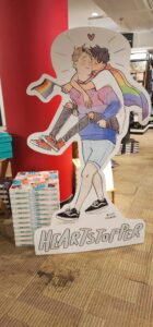 A cardboard cut out of Charlie and Nick from the webcomic Heartstopper. Using Pride Month, Nick is wearing a bisexual sweater while carrying Charlie piggy back, who is wearing a rainbow flag as a cape and holding a mini flag in his hand. The duo stand perched in front of two stacks of the first volume of Heartstopper and a bright red pillar used to separate sections of the floors.