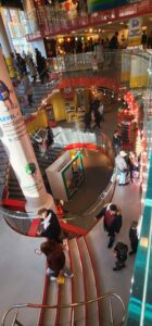 A large grey and red spiral staircase descends down three floors below street level. Each floor is cluttered with bright colors, lights, and a myriad of M&M merchandise.