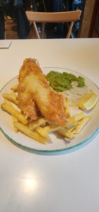 A singular golden piece of fried fish sits on a bed of yellow fries. Next to it is a puddle of tarter sauce and a pile of moss green mushy peas. All atop a white circular plate.