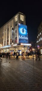 A large poster advertising Matilda sits attached at the front of the Cambridge Theatre, a large white, almost triangular theatre with large windows trailing the walls in either direction. 