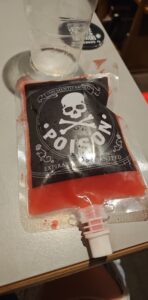 A Halloween themed, blood red cocktail inside an applesauce pouch like bag. The black label has a large skull and cross bones on it in white with POISON scrawled underneath.