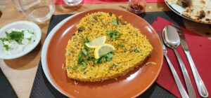 On an orange circular plate sits a triangular shaped mass of yellow rice with lamb chunks and cilantro on top. Two lemon wedges garnish the top.