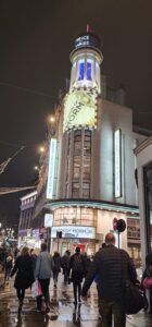 The historic Prince of Wales Theatre, the rounded corner hosts the large Book of Mormon poster and windows down beneath it. The marquee is lit in bright lights and more posters of the show. A large group of people walk towards the theatre at the bottom of the photo.