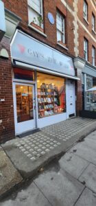A quaint little light blue exterior with “Gay’s the Word” scribbled on the overhead sign. Warm Light emits from the shop interior through the window, creating a warm atmosphere. A display of current and new books sits nicely organized in the large front window.
