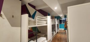 Rows of bunk beds pushed up against a painted, geometric purple, blue, and white wall. The far wall is flat blue and is occupied by a frosted window leading into the smoking area.