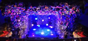 The stage for Matilda. The entire wall is covered in scrabble like tiles, each with a different letter on them. Swings with more scrabble tiles descend from the ceiling spelling out Matilda in rainbow colors. The whole stage is cast in a deep blue light.