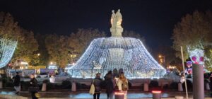 The large beige fountain sits in the center of the photo, white Christmas lights cascading down from the top to the bottom. Four girls stand at its base admiring it.