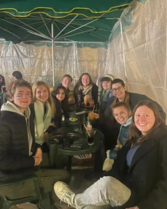 A group shot of 9 students, myself included, under a green umbrella and surrounding green plastic tables adorned with various drinks. Surrounding us is a semi-opaque white plastic wind blocker attached to the umbrellas. Everyone is dressed for winter in coats and scarves. Some students have the French flag on their face, made with face paint.