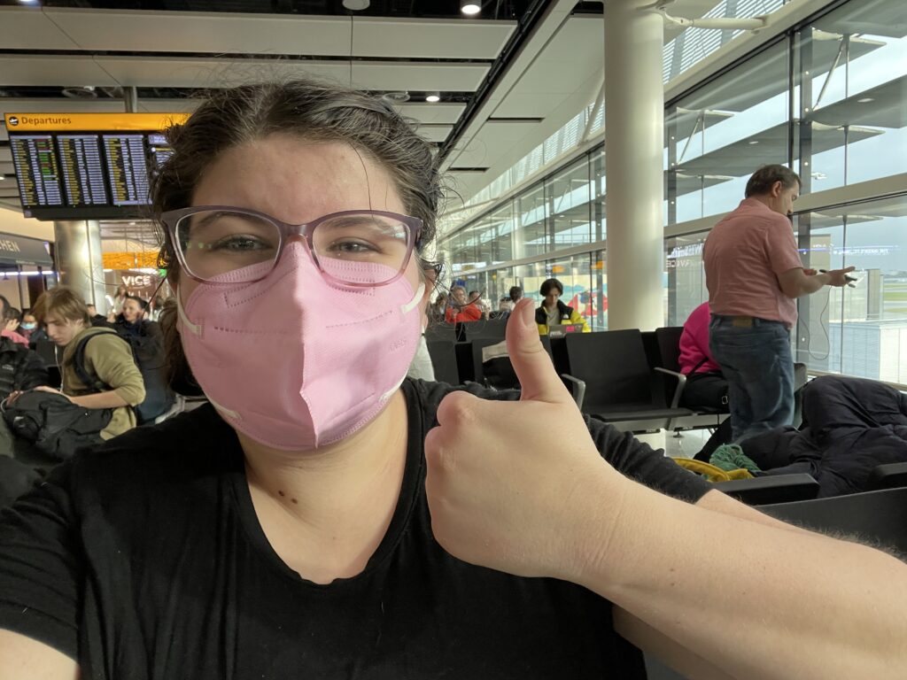 A young woman (Katie) wearing a pink KN95 mask and giving a thumbs up. She is wearing a black t-shirt and her hair is in two braids. She is sitting in an airport terminal