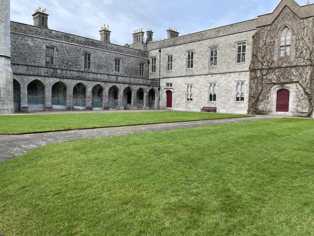 picture of an L-shaped building made of light grey stone with a grass courtyard in front of it