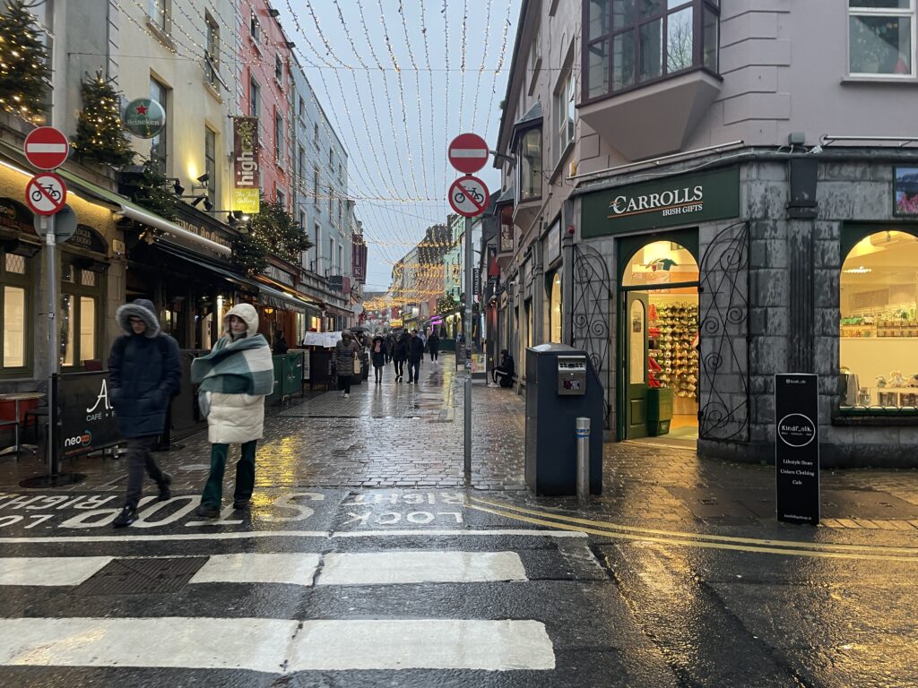 picture of a narrow street with buildings lining both sides. In between the buildings are strings of lights. It is raining and the ground is wet