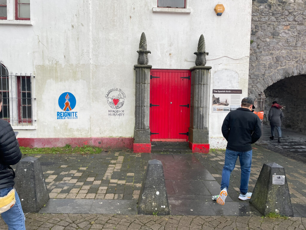 a white wall with stencil graffiti and a red door. One piece of stencil graffiti is a blue circle with two red hands clasped together with a red flame above them. Below is blue text that reads "reignite world freedom". The other is a circle with a black and white silhouette of the galway cathedral in the background and two black and white hands holding a red heart. Around the art are the words "Claddagh Watch, Heroes of Humanity". The wall also has a bright red door and merges with a dark grey stone arch towards the right of the photo