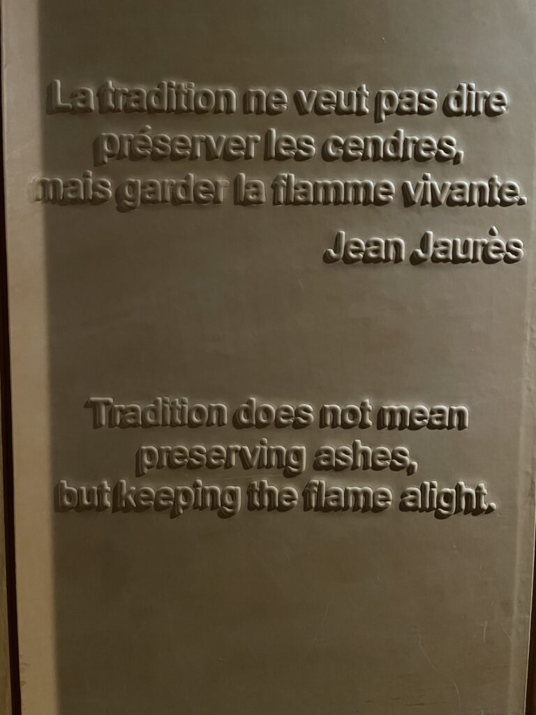 Text embossed on stone. The top passage, in French, reads "La tradition ne veut pas dire preserver les cendres, mais garder la flamme vivante.- Jean Jaures". The bottom passage is a translation of the top passage and reads "Tradition does not mean preserving ashes, but keeping the flame alight."
