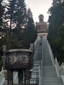View of the Tian Tan Buddha between trees to the left and right, from the bottom of the flight of stairs, around sunset. 