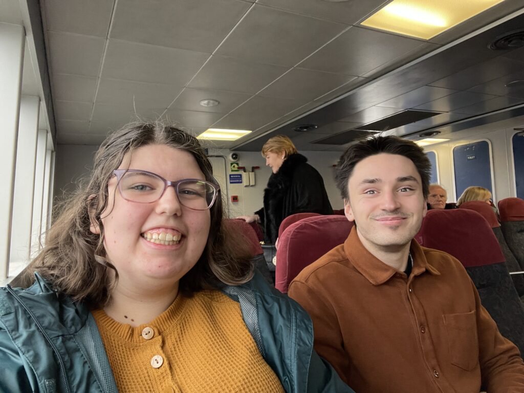 two people sitting on a ferry. The person on the left is a young woman wearing a teal jacket and a yellow shirt. The person on the right is a young man wearing a brown shirt