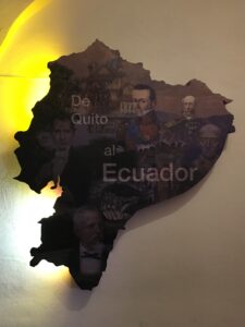 Pictured is a wooden carving of Ecuador's shape. The photo has a few historical figures painted onto the sign 