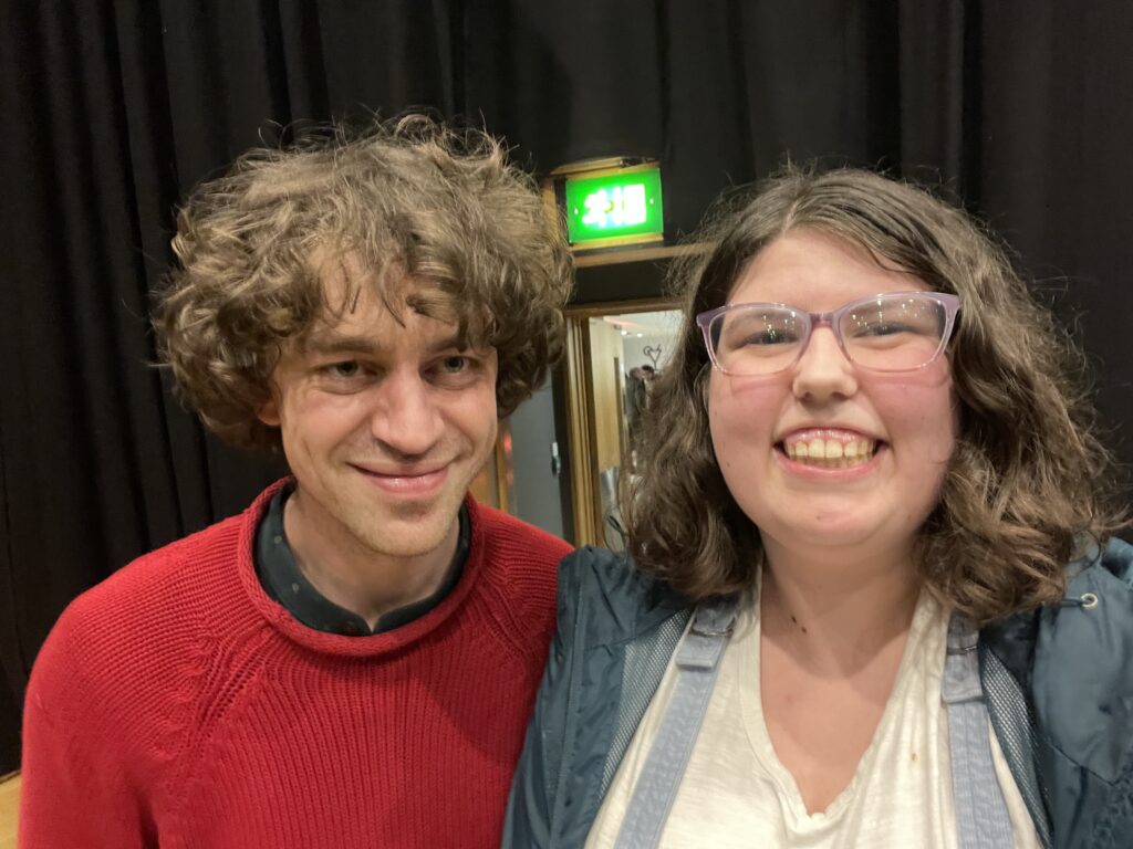 two people: a man in a red sweater with curly hair on the left, a girl wearing overalls and glasses on the right
