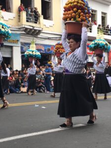 Pictured is a part of the Ambato parade. There are women wearing traditional clothing, walking down the street, and carrying flowerpots on their heads.