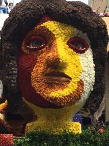 Pictured is part of an Ambato parade float. This float is a giant head that is made of brown, red, white, and yellow flowers. 