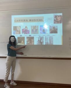 Pictured is me smiling and pointing at a slide show presentation that I made. On this slide is every one of Taylor Swift's albums covers in a timeline of their release dates