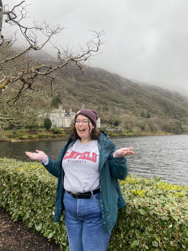 a girl wearing a grey "Linfield Wildcats" sweatshirt, a teal rain jacket, and a purple beanie in front of a castle