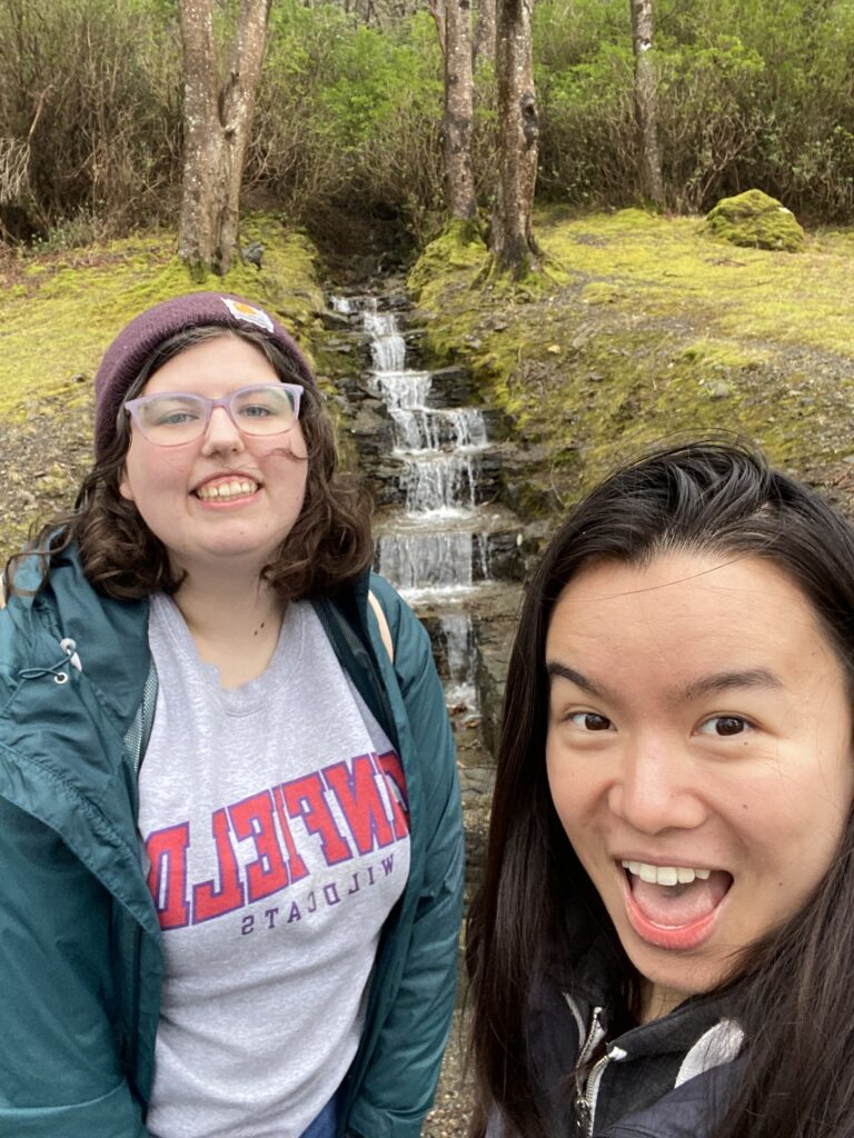 two girls in front of a waterfall. The one on the left is wearing a grey "Linfield Wildcats" sweatshirt, a teal rain jacket, and a purple beanie. The one on the right is wearing a grey rain jacket