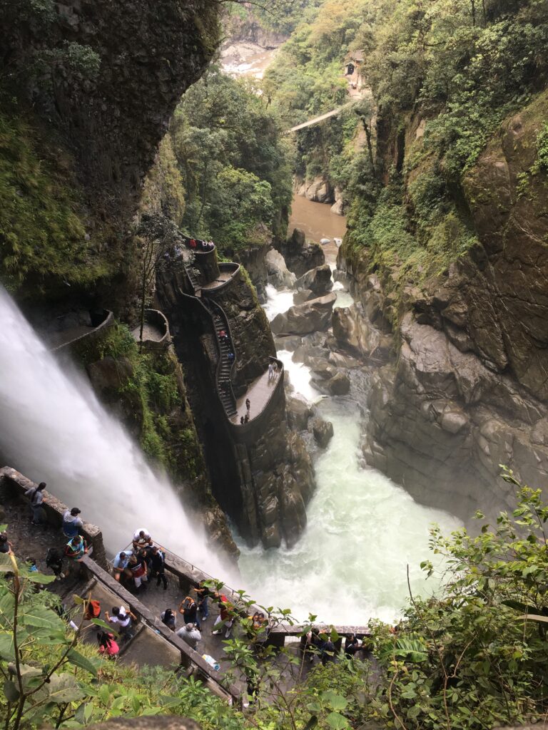 Pictured is the side view of a waterfall. Surrounding the waterfall is various greenery, naturally-carved rocks. and a below water outlet for the waterfall. 